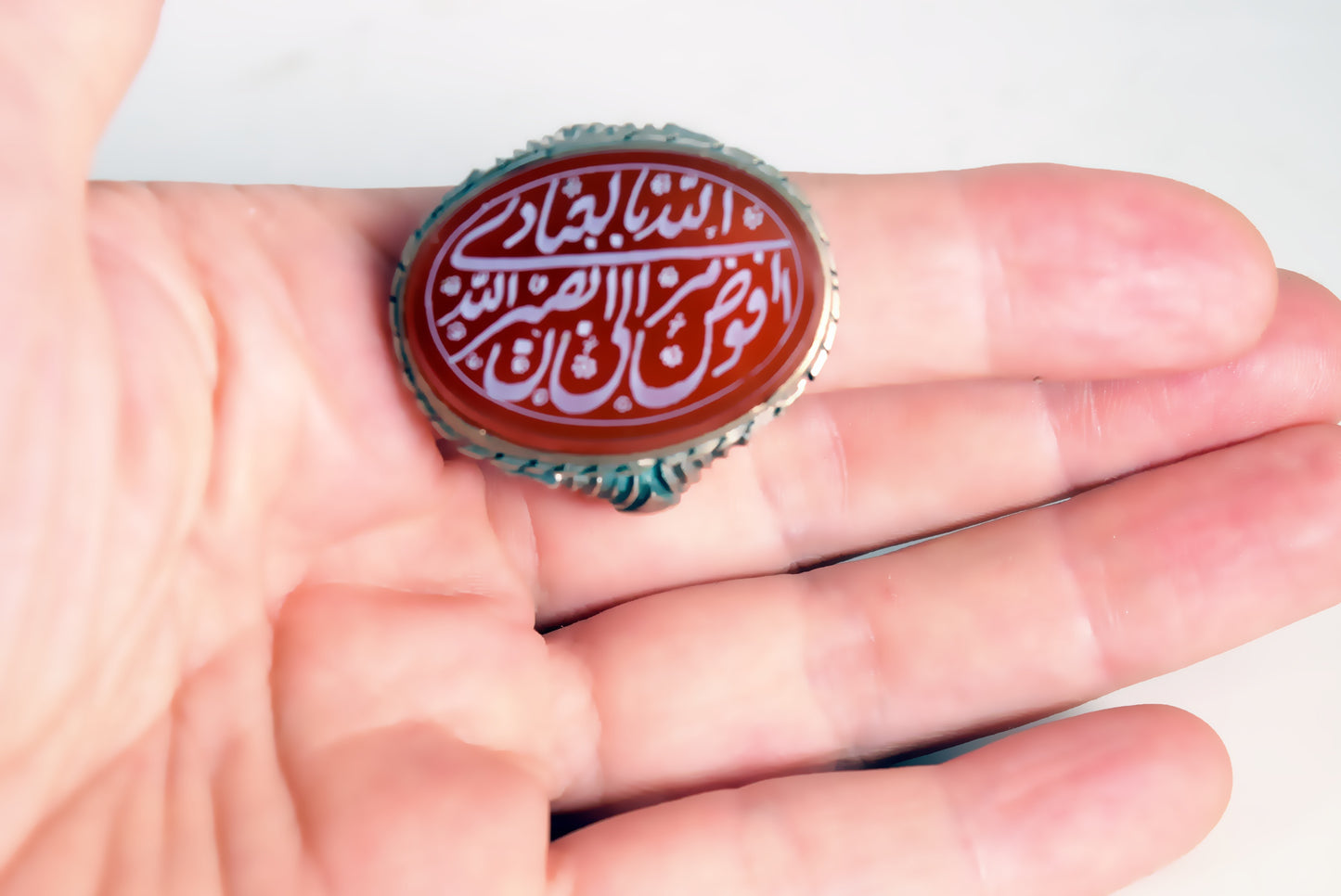 A hand holding a silver ring with an oval red cabochon featuring white Arabic calligraphy.