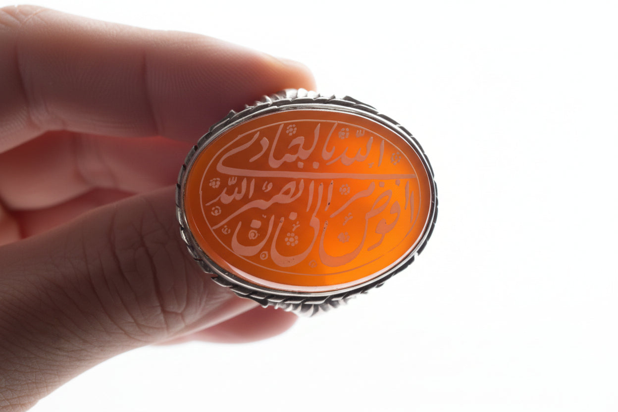A hand holding a silver ring with an oval orange cabochon featuring Arabic calligraphy.
