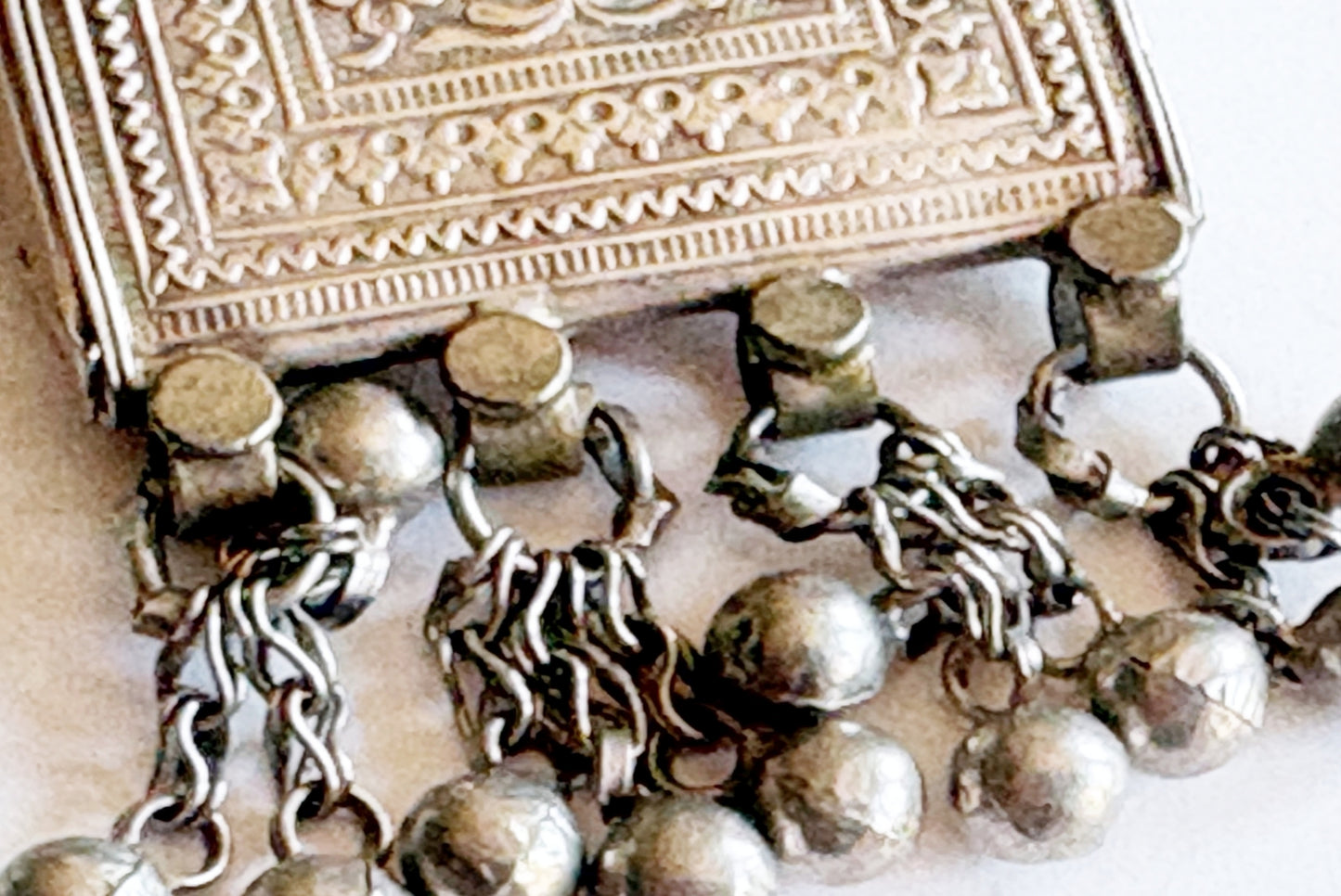 A close-up of an ornate silver-toned metal jewelry piece with an embossed design and dangling chain tassels ending in round beads.