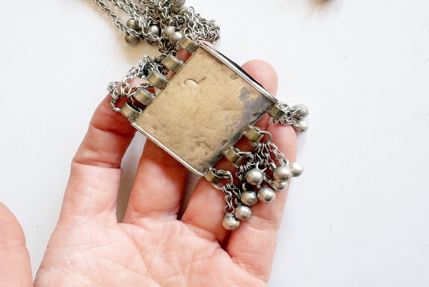 A hand holding a silver pendant necklace with a rectangular hammered metal center and dangling beaded chains, shown against a plain white background.