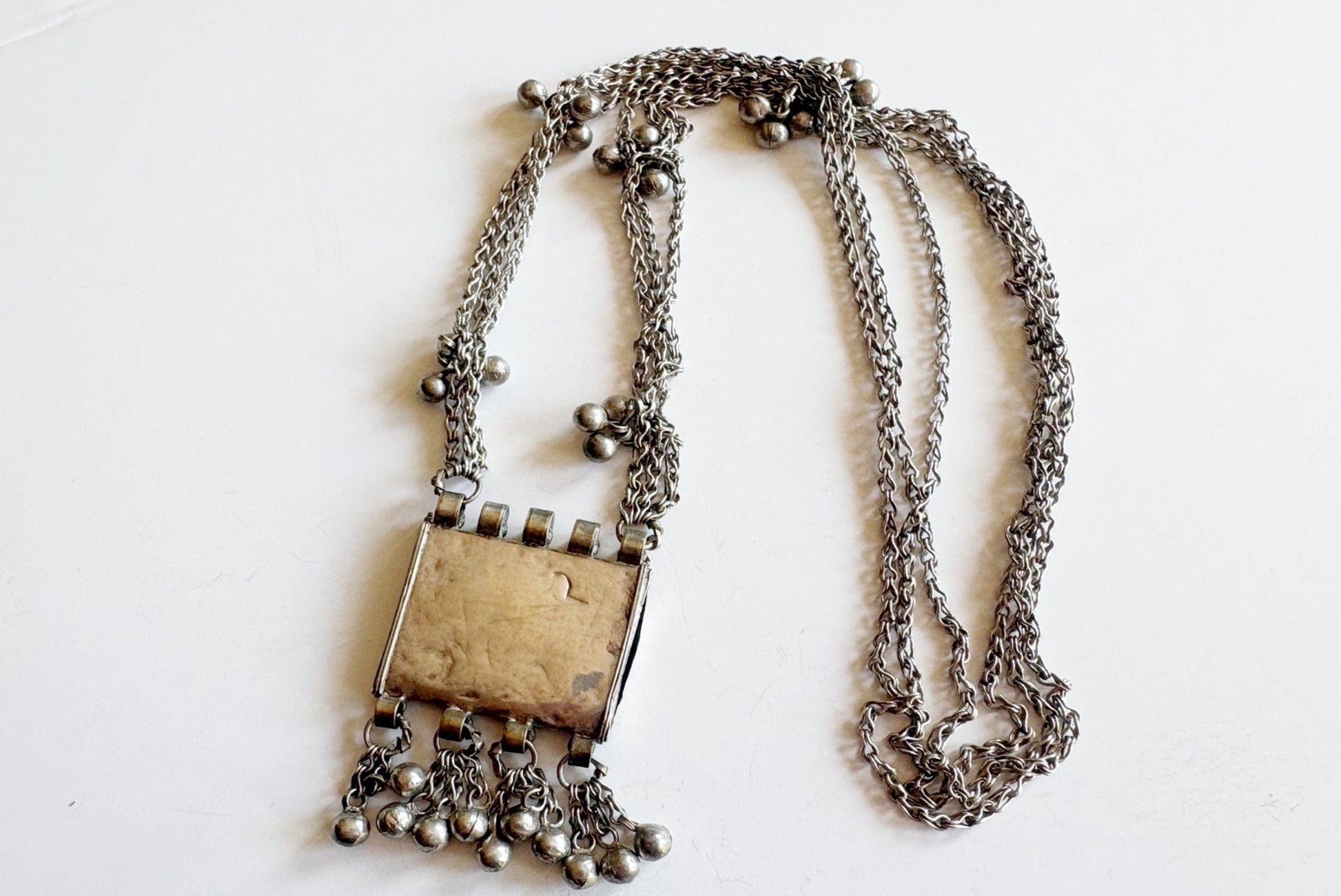 The back of a silver necklace featuring a rectangular pendant, surrounded by dangling chains and small silver beads, displayed on a plain white background.