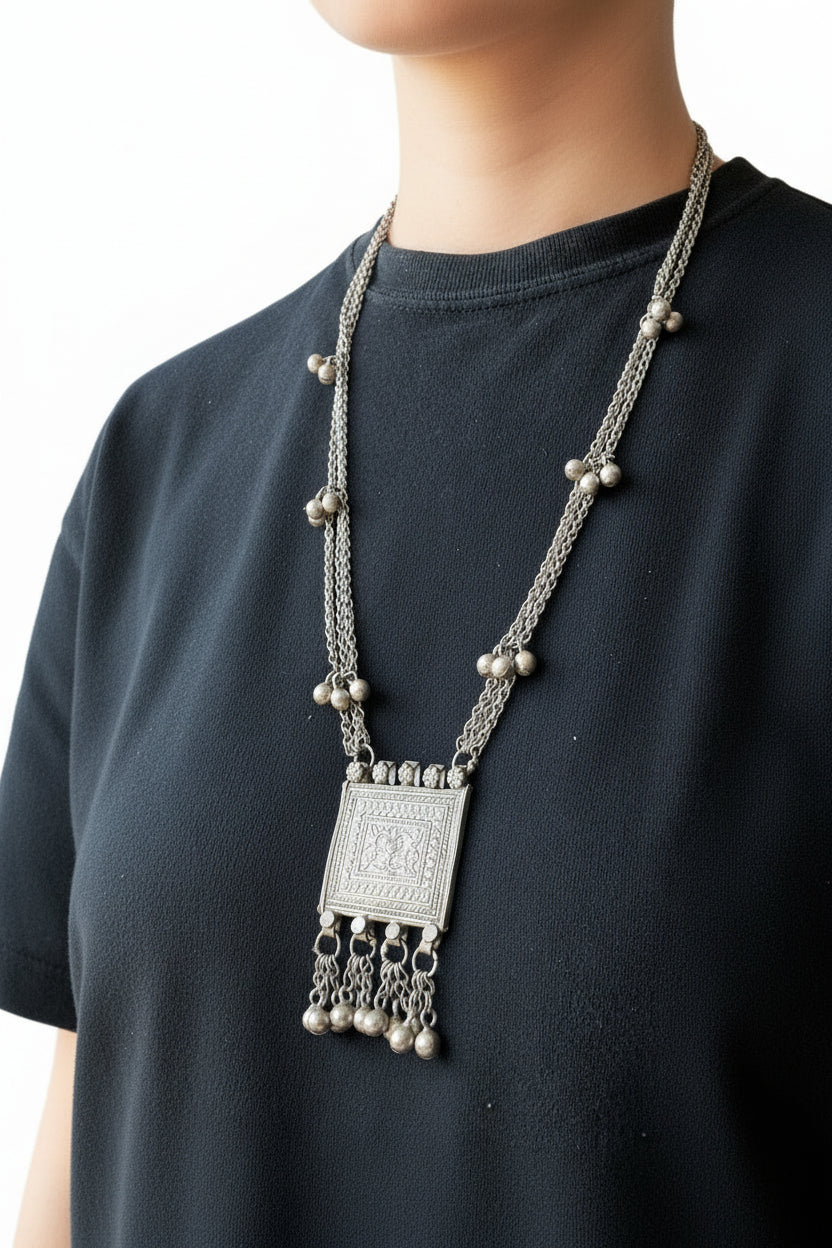 A person wearing a long silver-toned chain necklace with a square engraved pendant and dangling bell charms, layered over a black t-shirt.
