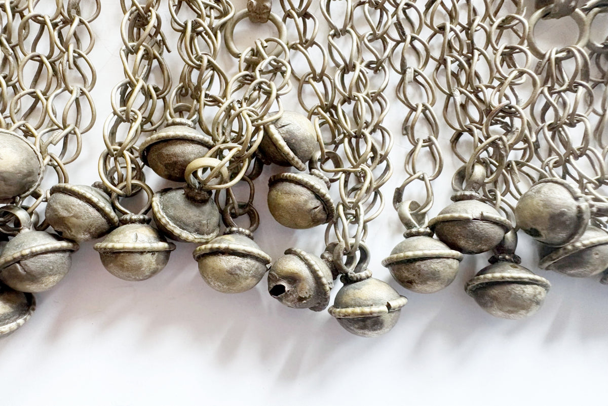 A close-up of multiple silver-toned bell shaped pendants hanging from chain links, arranged side by side on a plain white background.
