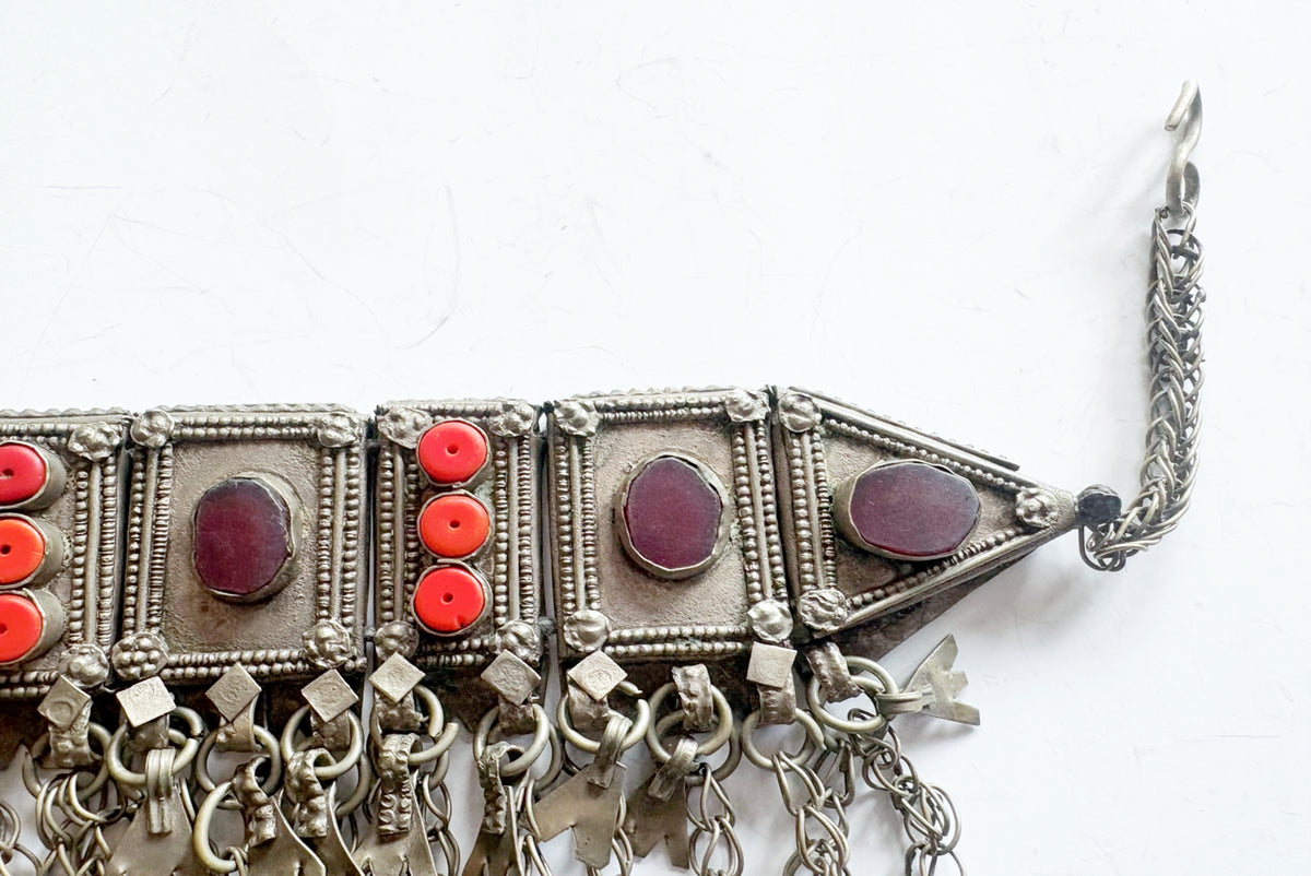 An ornate silver-toned traditional necklace with rectangular panels, dangling chains, and red gemstone accents on a plain white background.
