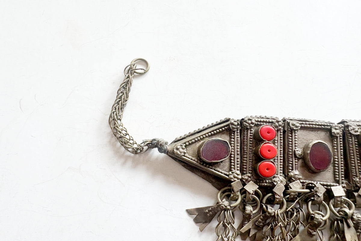 A close-up of a silver-toned traditional necklace with geometric metalwork, dangling charms, and three red circular beads on a white background.