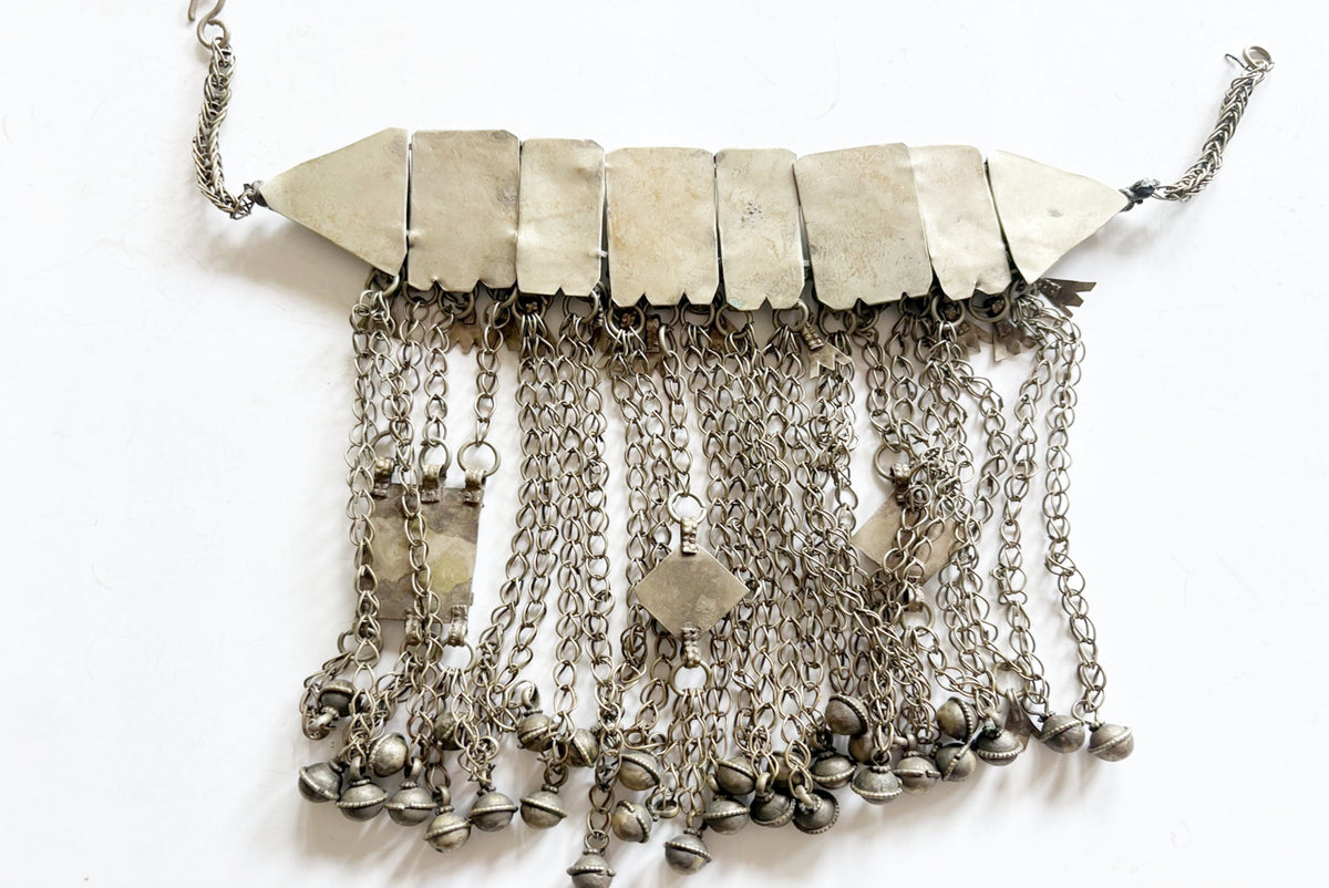 An antique-style silver-toned metal collar necklace featuring a horizontal bar of pointed triangular plates with multiple dangling chains and small bell charms, shown on a plain white background.