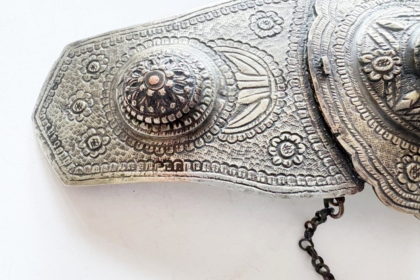 An ornate metal belt buckle with intricate floral and geometric engravings, featuring a decorative central medallion and a dark metal chain.