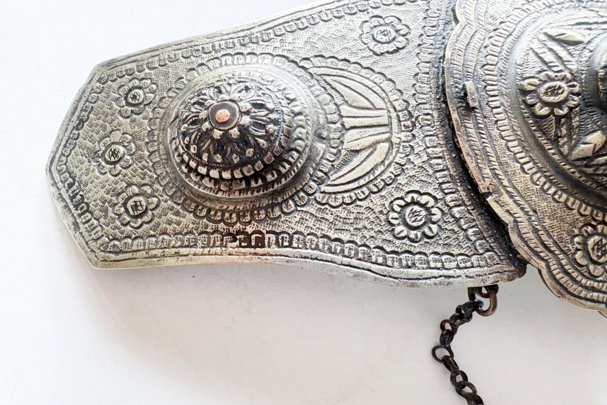 An ornate metal belt buckle with intricate floral and geometric engravings, featuring a decorative central medallion and a dark metal chain.