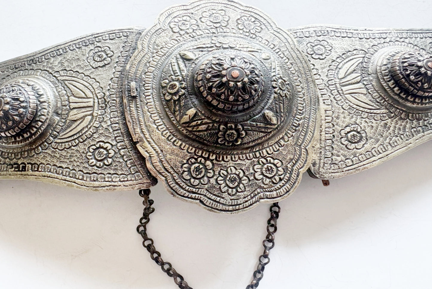 An ornate metal belt with intricate embossed floral and geometric patterns and a short hanging chain, shown against a plain white background.