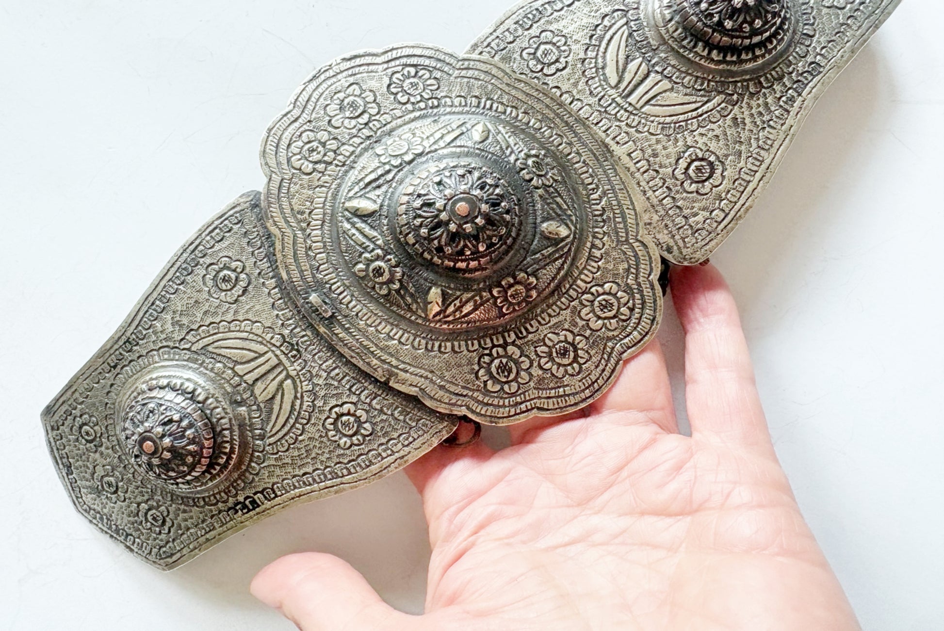 An ornate metal belt with intricate floral engravings and three large decorative conchos, held by a hand against a plain white background.