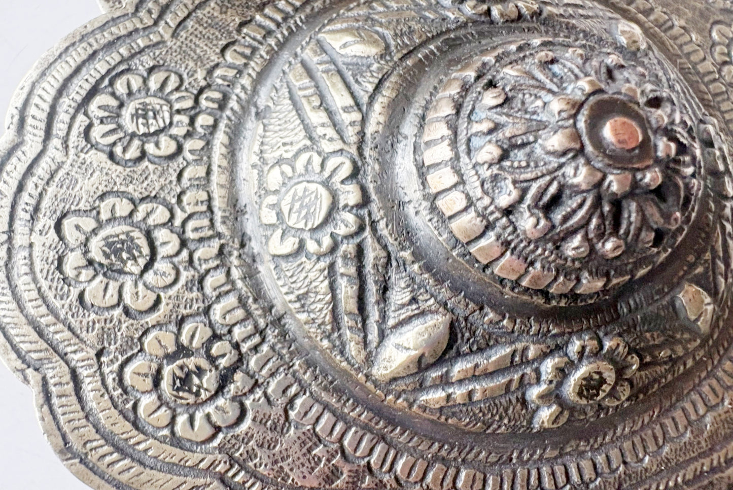 Close-up of an ornate circular metallic decorative belt buckle with embossed floral and geometric patterns, showing a silver-toned surface with darker aged areas.