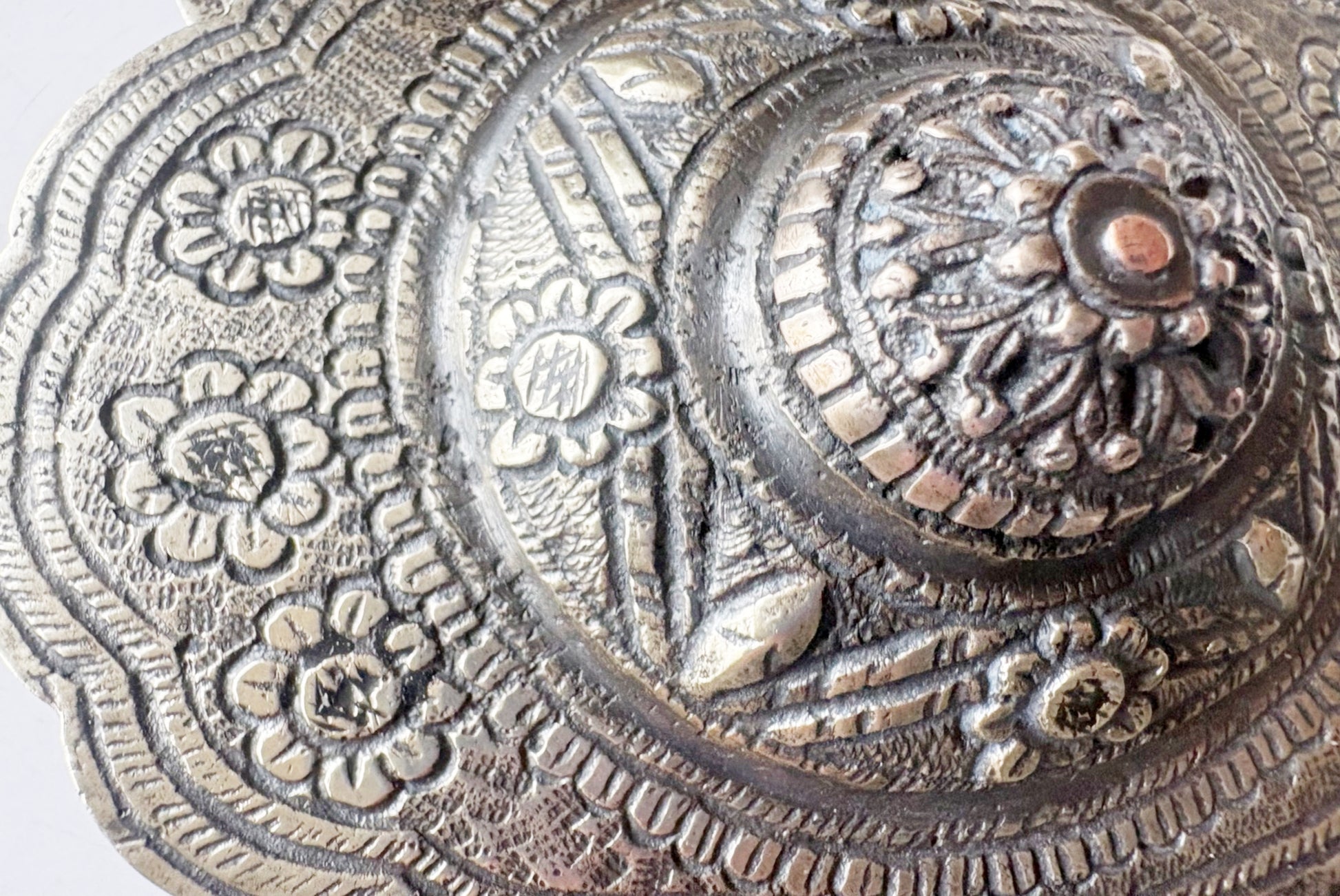 Close-up of an ornate circular metallic decorative belt buckle with embossed floral and geometric patterns, showing a silver-toned surface with darker aged areas.