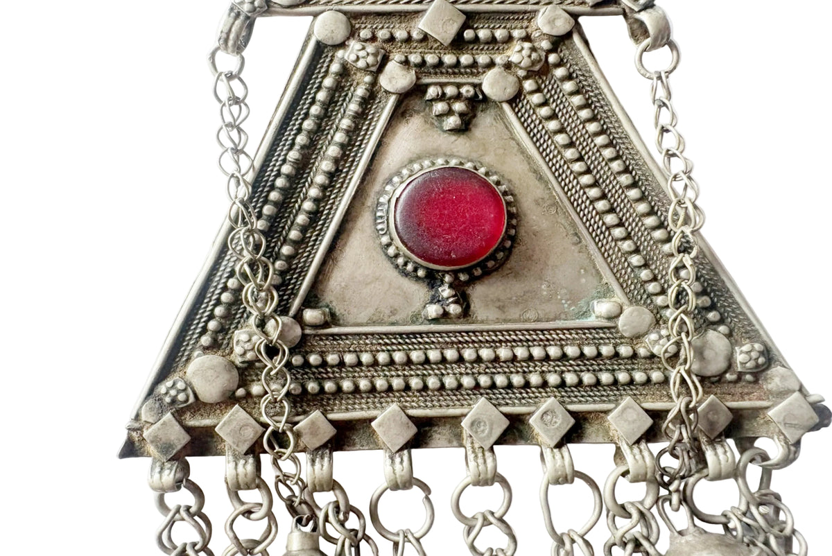 A close-up of a silver-toned triangular pendant or wall hanging with intricate beadwork, chain drapes, and a central round red stone on a plain white background.
