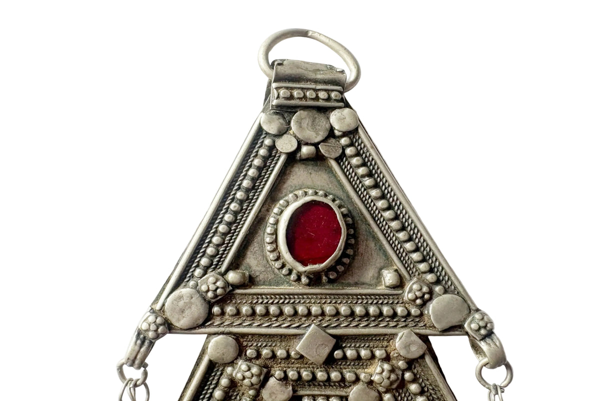An ornate triangular silver-toned pendant featuring intricate beadwork, filigree details, and a central oval red gemstone, shown against a plain white background.