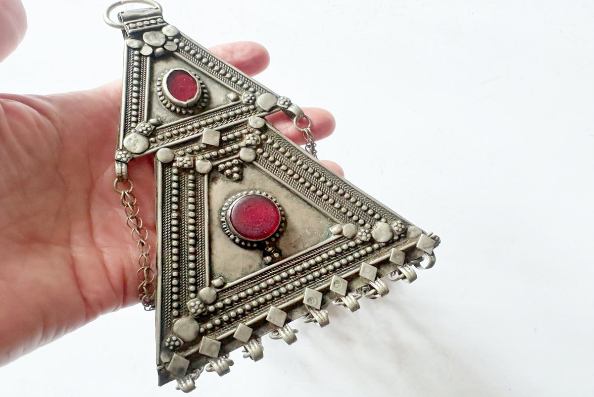 A hand holding a large, ornate silver-toned triangular pendant with two red circular stones and intricate geometric detailing, set against a plain white background.