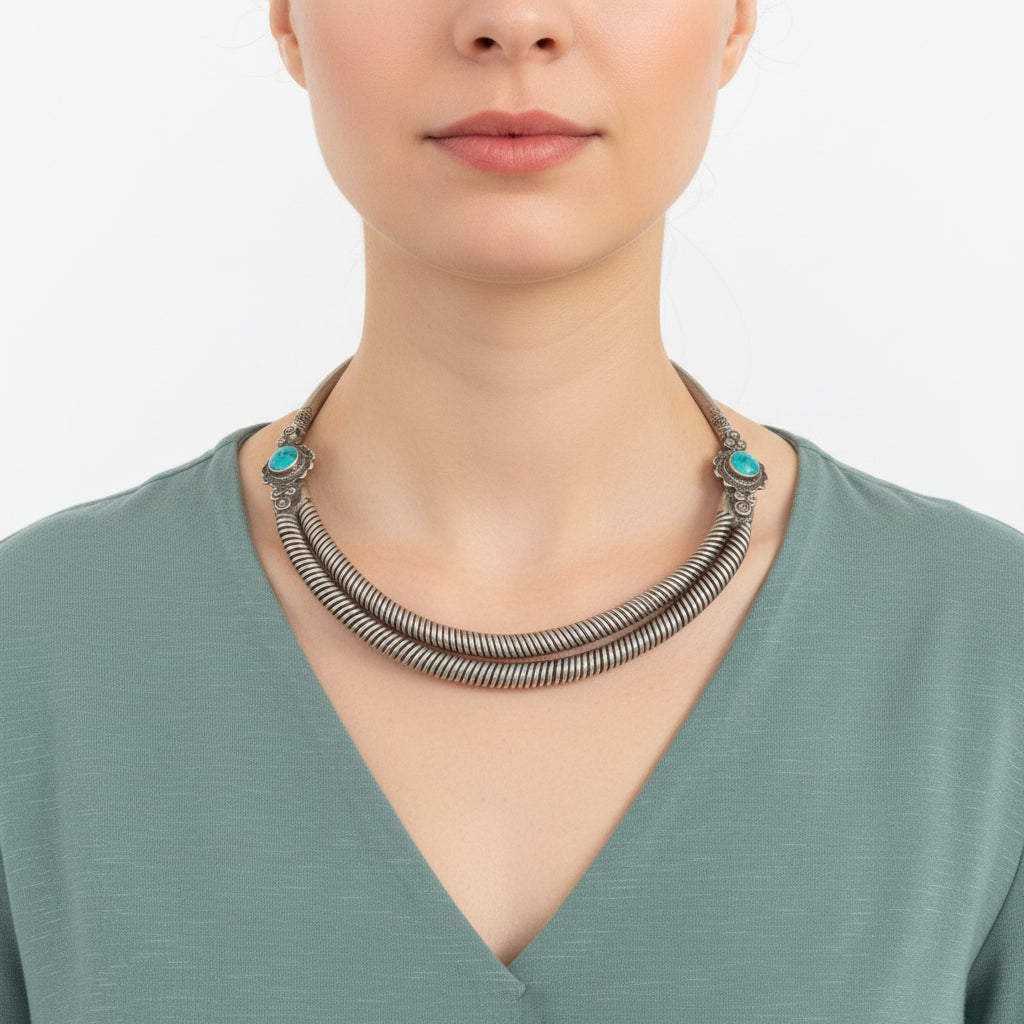 A close-up of a person wearing a chunky silver-toned double-strand coiled necklace with turquoise stone accents at the ends, paired with a sage green top.