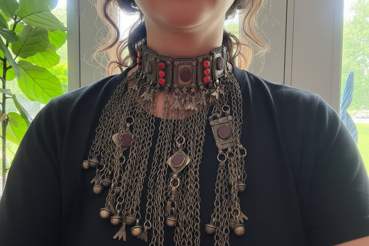 A close-up of a person wearing an ornate antique-style silver-toned choker necklace with red gemstone accents and multiple dangling chain elements over a black top.