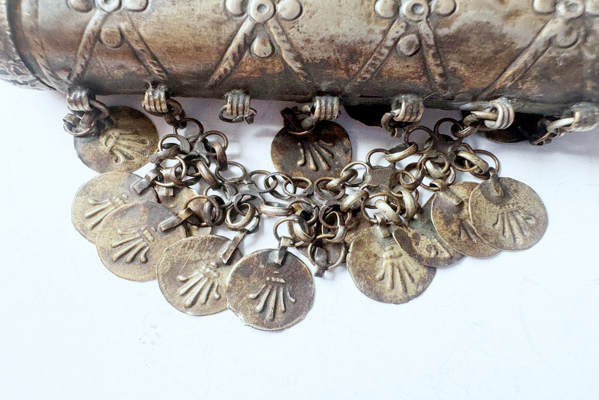 A close-up of a vintage silver-toned metal textured cylindrical pendant with hanging coin charms.