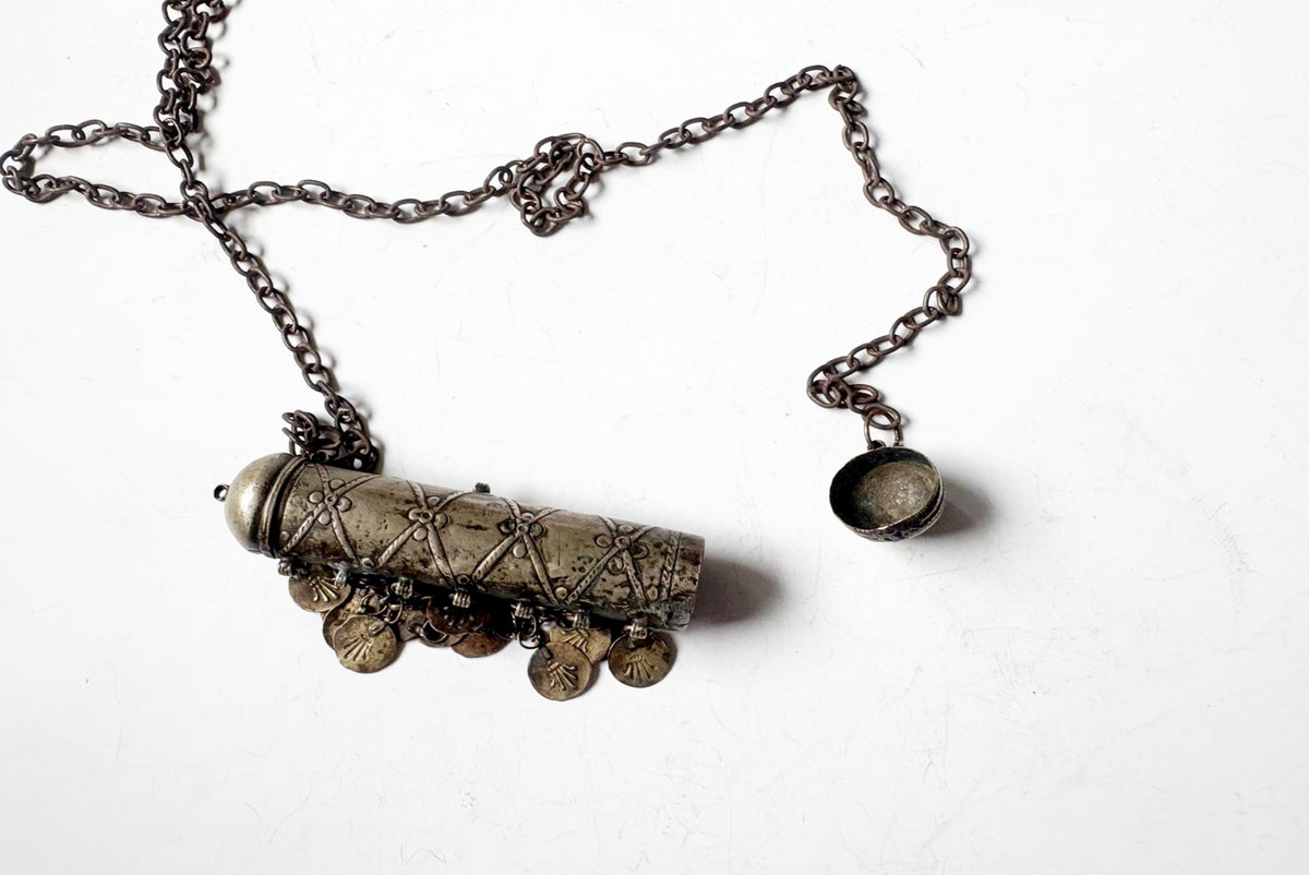An antique-style silver-toned necklace featuring a cylindrical pendant with intricate geometric patterns, dangling coin charms, and a separate small oval charm on a dark chain, displayed on a white background.
