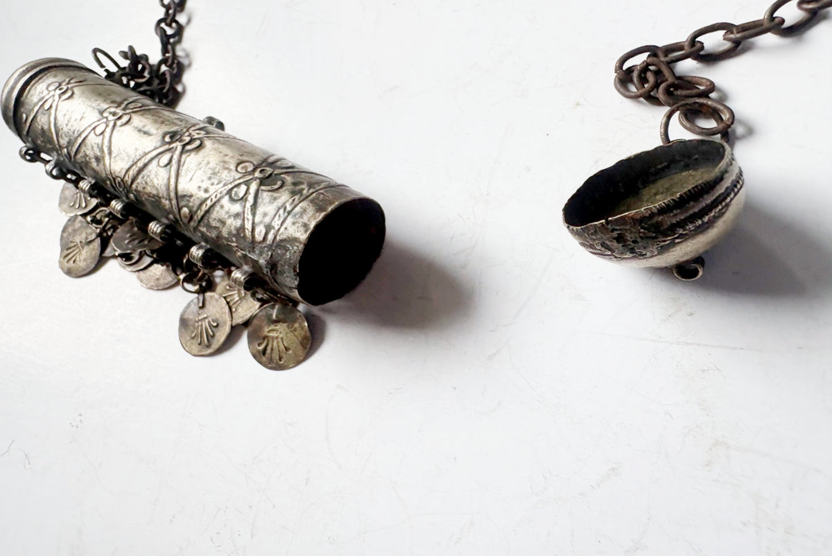 An antique-style silver-toned pendant necklace shown in two pieces on a white surface: a cylindrical capsule pendant with dangling coin charms on the left, and a small dome-shaped pendant on the right.