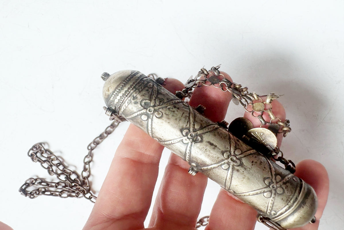 A close-up of a hand holding a vintage silver-toned cylindrical pendant or locket with engraved floral patterns and a chain, against a plain white background.