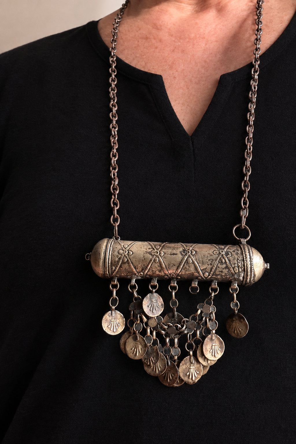 A close-up of a person wearing a bohemian-style bronze and silver pendant necklace. The necklace features a textured cylindrical bar with dangling coin charms, worn over a black top.