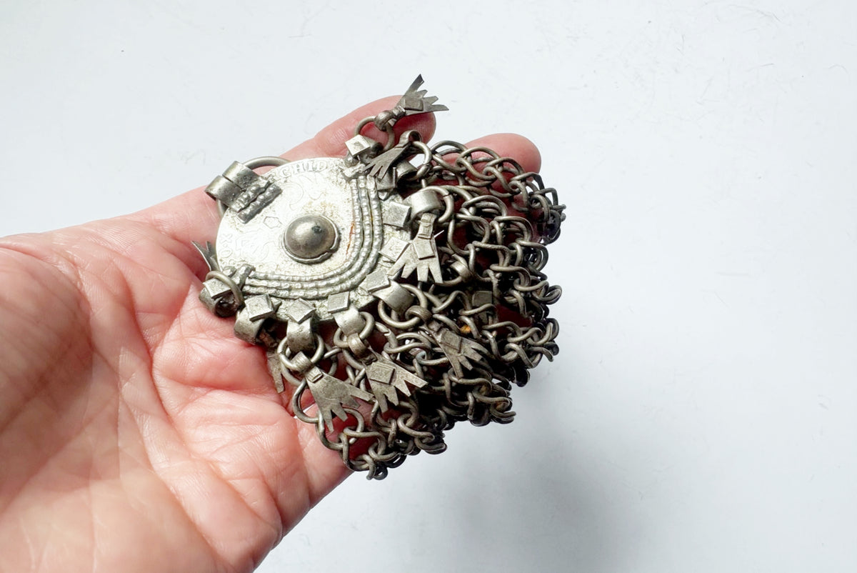A hand holding a silver-toned traditional Bedouin pendant with a circular Maria Theresa coin central bead, and dangling chain-like elements against a plain light background.