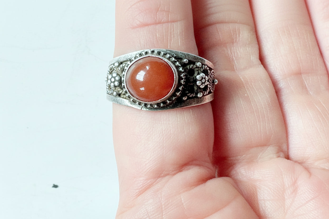 A silver ring with an orange stone, worn on a person's finger against a light background.