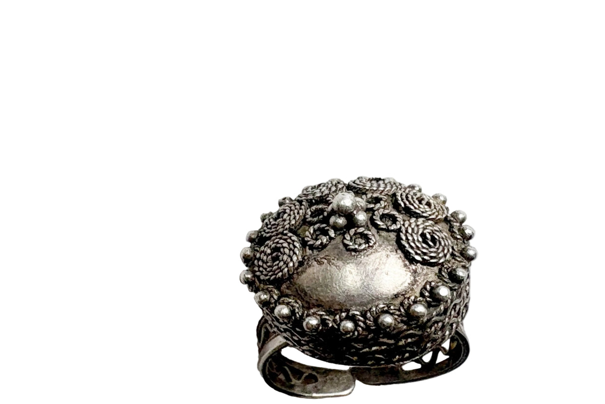 An ornate vintage sterling silver ring with a domed lid decorated with intricate floral scrollwork and beaded borders, resting on a white surface.