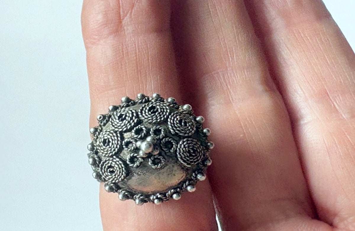 An ornate silver ring with intricate floral and spiral filigree patterns, shown worn on a person's finger against a light gray background.